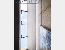 Beton-Innenraum mit hohen, glatten Wänden, einem großen rechteckigen Deckenfenster, das den Himmel zeigt, und Metallstreben im oberen Bereich. Auf einer der Wände ist ein abstraktes Kunstwerk sichtbar.