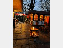 Ein Außenbereich mit einer kleinen Hütte, bezeichnet als „Rodelhütte“, mit beleuchteten Fenstern und einer roten Leuchtschrift. Im Vordergrund steht eine brennende Feuerschale auf einem gepflasterten Weg, umgeben von Töpfen und winterlicher Vegetation, während hinter der Hütte Bäume im Abenddämmerungslicht sichtbar sind.