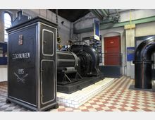 Innenansicht eines industriellen Museumsraums mit historischen Maschinen, darunter ein großes schwarzes Gehäuse mit der Aufschrift „Zeichnungen 1893“ und eine maschinelle Anlage mit Rohren und Manometern. Die Umgebung ist schlicht, mit Fliesenboden in Rautenmuster, weißen Wänden und einer roten Tür im Hintergrund.