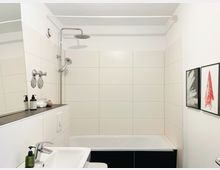 Modernes Badezimmer mit weiß gefliester Wand, Badewanne mit integrierter Regendusche und Handbrause. Im Vordergrund ein Waschbecken mit Spendern, links ein großer Spiegel und rechts zwei gerahmte Wandbilder.