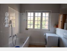 Helles Badezimmer mit weiß gefliesten Wänden, Badewanne mit Handbrause links und einem großen, sprossigen Fenster in der Mitte. Rechts stehen eine Waschmaschine unter einer Ablage sowie ein hölzerner Wandschrank; der Boden ist gefliest.