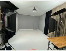 Ein Fotostudio mit grauen Wänden und einem weißen Boden, der mit orangefarbenen Markierungen versehen ist. Zwei große Softboxen zur Beleuchtung stehen auf Stativen, und an der rechten Seite befindet sich ein Holztisch; lichtundurchlässige Vorhänge bedecken die Wände.