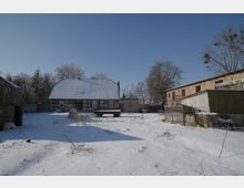 Verschneiter Hof eines ländlichen Anwesens mit einem Fachwerkhaus und schneebedecktem Satteldach in der Mitte, umgeben von Nebengebäuden und Zäunen. Im Vordergrund liegt eine offene, schneebedeckte Fläche mit verstreuten Pflanzenresten und Spuren, dahinter stehen kahle Bäume vor klarem Himmel.
