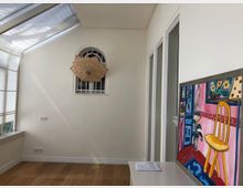 Ein heller Innenraum mit Holzboden und weißen Wänden. Links befinden sich große Fenster mit halbtransparenten Vorhängen, rechts ein Gemälde mit buntem Motiv auf einer weißen Kommode. Eine kleine, gewölbte Fensteröffnung mit einem dekorativen Papierfächer ist an der hinteren Wand zu sehen.