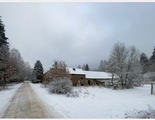 Ländliche Winterlandschaft mit einem schneebedeckten Weg, der an einem Backsteinhaus mit weißem Dach vorbeiführt. Umgeben von kahlen Bäumen und Büschen, die mit Schnee bedeckt sind.