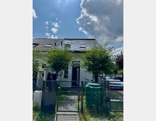 Reihenhaus mit weißer Fassade und dunklem Schrägdach, ausgestattet mit Dachfenstern. Vor dem Haus ein kleiner Garten mit zwei jungen Bäumen, gepflastertem Weg und Metallzaun; im Vordergrund Mülltonnen und ein Eingangstor.