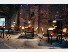 Industrie-inspirierter Café- oder Restaurantinnenraum mit unverputzten Ziegelwänden, Betonboden und einfachem Mobiliar aus Holzstühlen und Holztischen. Über jedem Tisch hängt eine einzelne Lampe, und auf den Tischen stehen kleine Blumenväschen.