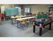 Ein Klassenraum mit gemischter Nutzung, ausgestattet mit Schultischen, Holzstühlen, einer grünen Tafel mit Zeichnungen, Regalen mit Spielsachen und Büchern sowie einem Tischkicker im Vordergrund. Die Wände sind teils aus Holzpaneelen, teils aus gelbem Mauerwerk.