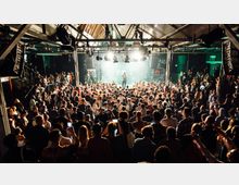 Eine große Menschenmenge versammelt sich in einem Konzertsaal mit industriellem Design, einschließlich einer metallenen Decke und sichtbaren Stahlträgern. Im Hintergrund beleuchtet eine Bühne einen Künstler mit Mikrofon, umgeben von Scheinwerfern und Graffiti-geschmückten Wänden.