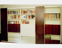 Große, wandfüllende Einbauregale in Weiß mit dunkelbraunen Holzschiebetüren, teils geöffnet und mit Büchern sowie Aktenordnern gefüllt. Unten befinden sich geschlossene Schrankfächer und Schubladen, oben mehrere offene Regalböden.