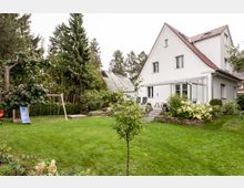 Ein Einfamilienhaus mit weißer Fassade und einem steilen Satteldach, umgeben von einem gepflegten Garten mit Rasenfläche, Sträuchern und Bäumen. Im Garten befinden sich ein Spielgerüst mit Rutsche und Holzschaukel sowie eine Terrasse mit Sitzmöbeln.