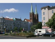 Straßenszene in einer Stadt mit historischen Gebäuden, einer Apotheke und einer großen Backsteinkirche mit hohen, grünen Türmen im Hintergrund. Mehrere Autos und Menschen sind sichtbar, sowie eine Straßenbahn-Haltestelle und Verkehrsstraßenschilder.