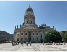 Der Französische Dom auf dem Gendarmenmarkt in Berlin, ein barockes Gebäude mit einem großen Kuppelturm und Statuen auf dem Dach. Im Vordergrund befindet sich ein gepflasterter Platz mit Menschen, im Hintergrund modernes Stadtgebäude und Bäume.