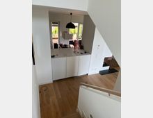 Blick in einen modernen, offenen Wohnbereich mit heller Küche und Holzfußboden. Die Küche verfügt über eine weiße Arbeitsplatte und einen schwarzen Hängeleuchter, im Hintergrund sind große Fenster mit Blick auf eine Terrasse und Grünanlage erkennbar.