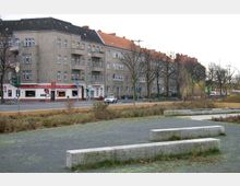 Straßenszene mit mehrstöckigen Wohnhäusern im Hintergrund, deren Fassade teils grau und teils mit roten Ziegeldächern gestaltet ist. Im Vordergrund befindet sich eine Grünfläche mit gepflasterten Wegen und steinernen Sitzbänken, daneben eine Hauptstraße mit einer Ampel und einem kleinen Geschäftsbetrieb.