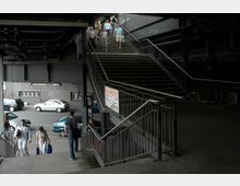 Eine Metalltreppe unter einer erhöhten Brückenkonstruktion, die zu einer Straße mit geparkten Autos führt. Im Hintergrund sind eine Backsteinwand mit Fenstern und mehrere Personen auf der Treppe zu sehen.