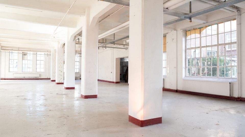 Heller, leerer Industrie- oder Loft-Innenraum mit glattem Betonboden und mehreren weißen Tragstützen mit roten Sockeln. Große Sprossenfenster lassen viel Tageslicht herein; an der Decke sind sichtbare Rohrleitungen und Kabeltrassen.