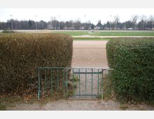 Ein kleiner Metallzaun mit einer geschlossenen, grünen Gartentür trennt einen Weg von einem Sportplatz mit Laufbahn und Rasenfläche. Im Hintergrund sind kahler Baumbestand, Flutlichtmasten und Häuser zu sehen.