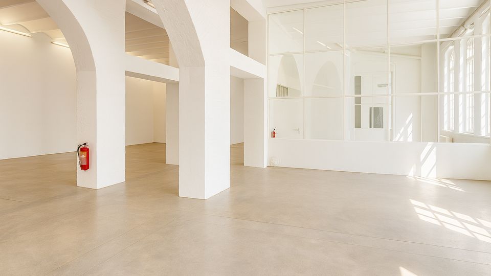 Heller, leerer Innenraum mit weißen Säulen und Rundbögen, glattem Betonboden und Deckenleuchten. Rechts trennt eine große Glaswand mit Sprossen einen Bereich ab; daneben fallen durch hohe Fensterfronten Tageslicht und Schatten auf den Boden.