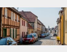 Schmale gepflasterte Straße in einer Altstadt mit anliegenden Fachwerkhäusern und farbigen Fassaden; am Straßenrand parken mehrere Autos, und im Hintergrund sind weitere Häuser und Bäume zu sehen.