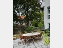 Ein Garten mit einer gepflasterten Terrasse, auf der ein ovaler Holztisch und mehrere Stühle mit dekorativen Lehnen stehen. Im Hintergrund befinden sich große Bäume, Büsche, ein Zaun und Wohnhäuser mit roten Dächern.