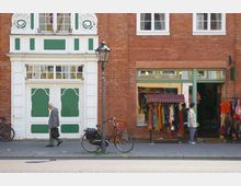 Eine Straßenansicht mit einer roten Backsteinfassade, links eine Tür mit grün-weißen dekorativen Elementen, rechts ein Schaufenster und ein Eingang zu einem Geschäft mit bunten Kleidungsstücken. Vor dem Gebäude stehen zwei Fahrräder und eine Straßenlaterne, während ein Mann vorbeigeht und eine Person am Laden steht.