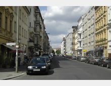 Straßenszene einer urbanen Wohngegend mit Reihen von mehrstöckigen Altbauhäusern, einigen mit dekorativen Fassaden. Parkende Autos säumen beide Straßenseiten, während Geschäftsflächen im Erdgeschoss teilweise sichtbar sind; Personen sind auf dem Gehweg zu erkennen, unter anderem neben Straßenschildern.