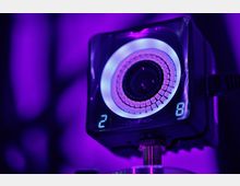 Nahaufnahme einer OptiTrack-Kamera mit einem leuchtenden LED-Ring um das Objektiv, dargestellt in violettem Licht. Im Hintergrund unscharfe, violette Lichtakzente.