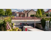 Straßenszene mit einer Betonbrücke im Vordergrund, darunter eine zweispurige Unterführung mit Autos und einem Radfahrer. Im Hintergrund sind Wohngebäude, ein gelbes Eckhaus und Bäume sichtbar.