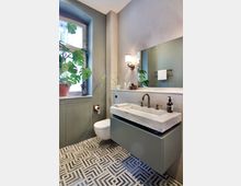 Modernes Badezimmer mit hellgrünen Wänden und schwarz-weiß gemustertem Boden. Ein Waschtisch mit Marmoroberfläche und goldfarbenem Armaturenset ist an der Wand montiert, daneben ein rundes WC, eine große rechteckige Spiegelwand sowie ein Fenster mit Pflanzen auf der Fensterbank.