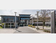 Modernes Verwaltungsgebäude mit großen Glasfassaden und horizontal angebrachten Lamellen zur Verschattung, flankiert von Bäumen und einer gepflasterten Gehwegfläche. Rechts im Bild steht eine Stele mit der Aufschrift „Kreishaus Teltow-Fläming“.