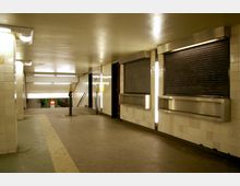 Unterirdischer Durchgang mit weißen Fliesenwänden und einem Boden aus Beton. An der rechten Seite sind geschlossene Rolltore und schmale Beleuchtungselemente angebracht, während im Hintergrund eine Treppe zu einer U-Bahn-Plattform führt.
