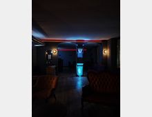 Dunkler Innenraum einer Lounge oder Bar mit gedämpfter Beleuchtung, sichtbar durch rote und blaue Lichtakzente an Decke und Säule. Möbel wie gepolsterte Sessel und Sofas, sowie eine dunkle Holzbodenfläche prägen das Ambiente.
