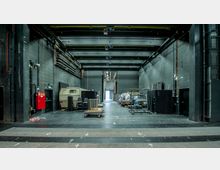 Blick auf den Hinterbereich einer Theaterbühne oder eines Veranstaltungsortes mit schwarzen Wänden und technischer Ausstattung wie Kabeln und Schienen an der Decke. Auf dem Boden stehen Requisiten, ein Anhänger, Fahrzeuge und gestapelte Stühle; im Hintergrund führt ein Gang zu einem hell beleuchteten Bereich.