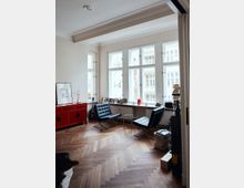 Wohnzimmer mit großen, weißen Sprossenfenstern, die viel natürliches Licht hereinlassen. Der Raum hat einen Fischgrät-Holzboden, eine rote Kommode, zwei schwarze Ledersessel mit einem runden Beistelltisch und eine Fensterbank mit Dekorationen und Bilderrahmen.