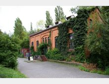 Backsteingebäude mit Rundbogenfenstern, teils von Efeu bewachsen, umgeben von Bäumen und Grünflächen. Ein gepflasterter Weg führt zu einem Eingang mit Treppenstufen, daneben eine Wand mit Graffiti und ein Aufsteller.