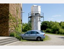 Ein bepflanztes Backsteingebäude mit Efeu an der Wand steht neben einem weiß gestrichenen Turm mit Metallkonstruktionen. Im Vordergrund ist ein blauer PKW auf einem gepflasterten Parkplatz zu sehen, im Hintergrund befinden sich Bäume und eine Wiese.