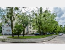 Straßenszene in einer Wohngegend mit einem mehrstöckigen beige-weißen Wohngebäude im Hintergrund, das kleine Fenster und einen modernen Glasblockeingang hat. Im Vordergrund stehen Bäume und Rasenflächen, während auf der Straße geparkte Autos zu sehen sind.
