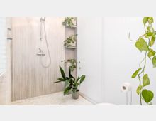 Modernes Badezimmer mit offener Dusche, beige gefliester Wand und mehreren Pflanzen in Töpfen, die die minimalistische Einrichtung ergänzen. Rechts im Bild sind eine Toilettenpapierhalterung und herabhängende Rankpflanzen zu sehen.