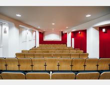 Ein kleiner Hörsaal mit Reihen von Holzsitzen, roten Wänden und einer weißen Decke. Im Vordergrund die Stuhlreihen, im Hintergrund eine weiße Leinwand an der roten Rückwand, beleuchtet durch Deckenstrahler.
