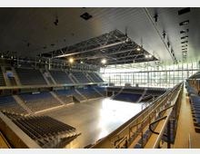 Innenansicht einer großen Mehrzweckhalle mit Sitzreihen auf mehreren Ebenen, einer hellen Fläche in der Mitte und einer verglasten Außenwand, die Tageslicht hereinlässt. Die Decke ist mit Metallträgern und Beleuchtungselementen ausgestattet.