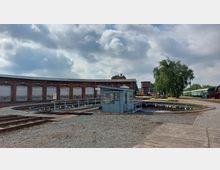 Halbrundes Bahnbetriebswerk mit roten Backsteintoren und einer Drehscheibe im Vordergrund. Rechts im Bild stehen historische Lokomotiven und grüne Waggons vor Bäumen und einem Wasserturm.