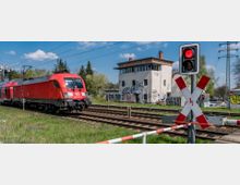 Ein roter Regionalzug der Deutschen Bahn fährt über Gleise an einem Bahnübergang mit einer geschlossenen Schranke und rotem Warnlicht. Im Hintergrund steht ein weißes, mit Graffiti versehenes Stellwerkgebäude, umgeben von Bäumen und Strommasten.