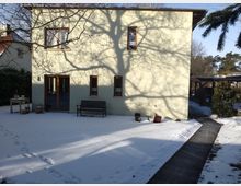 Zweistöckiges Wohnhaus mit hellgelber Fassade, umgeben von einem verschneiten Garten. Vor der Hauswand stehen eine Holzbank, Blumentöpfe und ein Tisch, während Schatten von Baumästen auf die Wand fallen; ein schmaler Holzweg führt durch den Schnee.