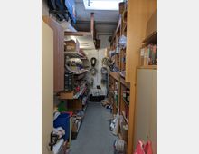 Ein schmaler Lagerraum mit Regalen auf beiden Seiten, gefüllt mit Kartons, Werkzeugen, Kabelrollen und verschiedenen Gegenständen. Im Hintergrund ist eine weiße Wand mit aufgehängten Kabeln und einer Kiste auf dem Boden sichtbar; eine Lichtkuppel sorgt für Tageslicht.