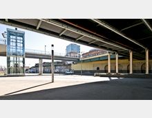 Außenansicht eines urbanen Bereichs unter einer Brücke mit sichtbaren Stahlträgern und Pfeilern. Im Hintergrund sind eine gläserne Aufzugsanlage, eine Hochbahnbrücke, mit Graffiti besprühte Wände und ein modernes Gebäude zu sehen.