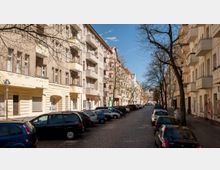 Straßenszene in einem Wohngebiet mit mehrstöckigen Altbauhäusern, verputzten Fassaden und Balkonen. Geparkte Autos säumen die gepflasterte Straße, während die kahlen Bäume entlang des Bürgersteigs dem Bild Struktur verleihen.