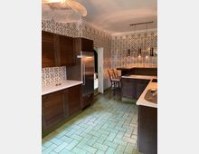 Küche mit dunklen Holzschränken, einer weißen Arbeitsplatte und gemusterten Fliesen an den Wänden mit grünem Pflanzenmotiv. Der Boden ist mit grünlichen Fliesen in Ziegeloptik ausgelegt, und eine Kochinsel mit Hockern sowie eine Edelstahlspüle sind sichtbar.