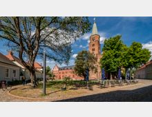 Historischer Platz mit einer Backsteinkirche und einem hohen Turm im Zentrum, umgeben von Bäumen und historischen Gebäuden mit Satteldächern. Im Vordergrund stehen Tische und Stühle eines Cafés auf einem gepflasterten Platz.