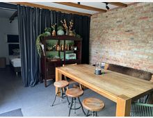 Ein rustikal eingerichteter Wohnraum mit einer freiliegenden Backsteinwand, einem Holztisch mit Hockern und einer Holzbank. Im Hintergrund steht ein dunkelbrauner Vitrinenschrank mit grünen Regalfächern, dekoriert mit Pflanzen und Keramikgegenständen; daneben ist ein abgetrennter Schlafbereich mit schwarzem Vorhang sichtbar.